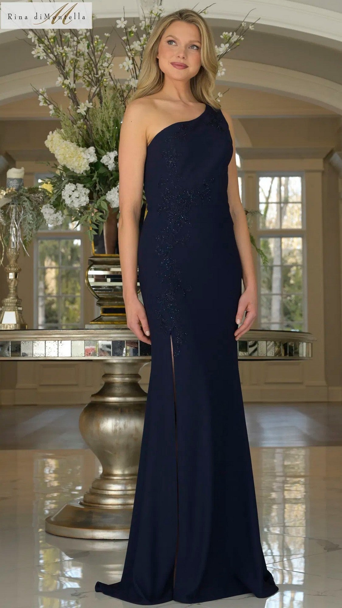 A woman stands indoors wearing the Rina Di Montella RD3240 long formal dress in navy, one-shouldered with subtle embellishments, posed in front of a large vase of white flowers.